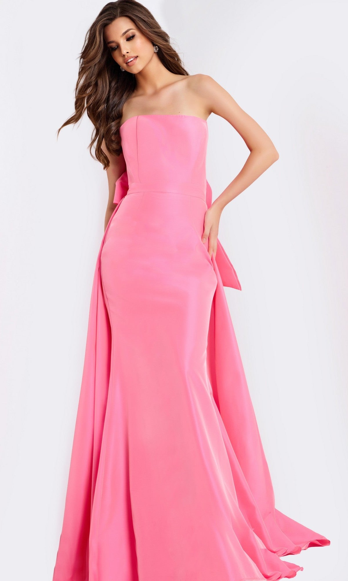 A woman models the Jovani 47495 long prom dress in bright pink, featuring a strapless fitted bodice and flared skirt, as she poses gracefully against a plain light background.